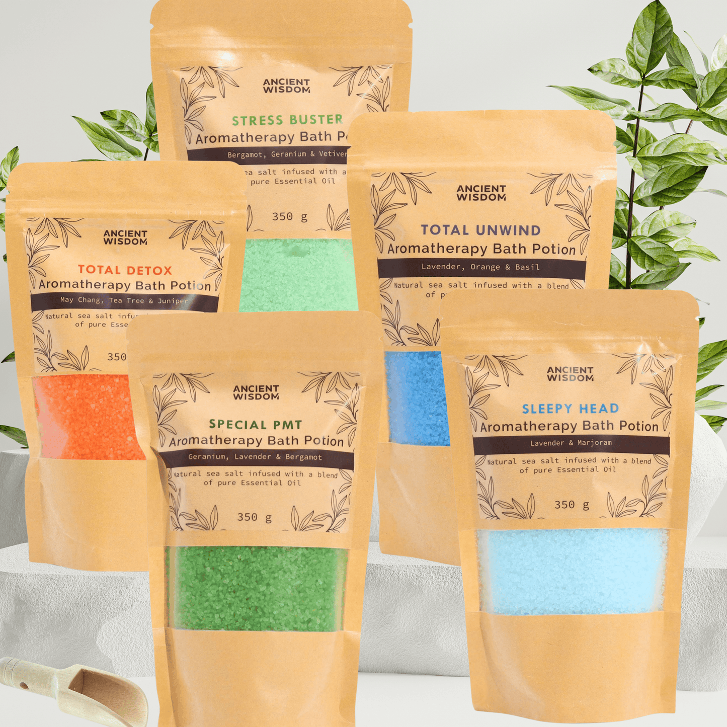 Five Kraft bags with aromatherapy bath salts in them. Each bag has a different colour bath salt in them and standing on a marble stone platform with a green plant in the background and a small wooden scoop infront of the bags