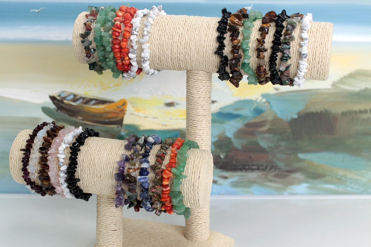 Chipstone bracelets of different colours hanging on a display stand in front of a painting of the seaside and and a small boat