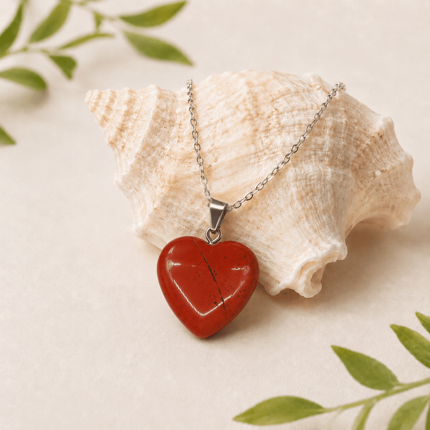 Heart-shaped red stone pendant on a silver chain with a shell and green leaves in the background