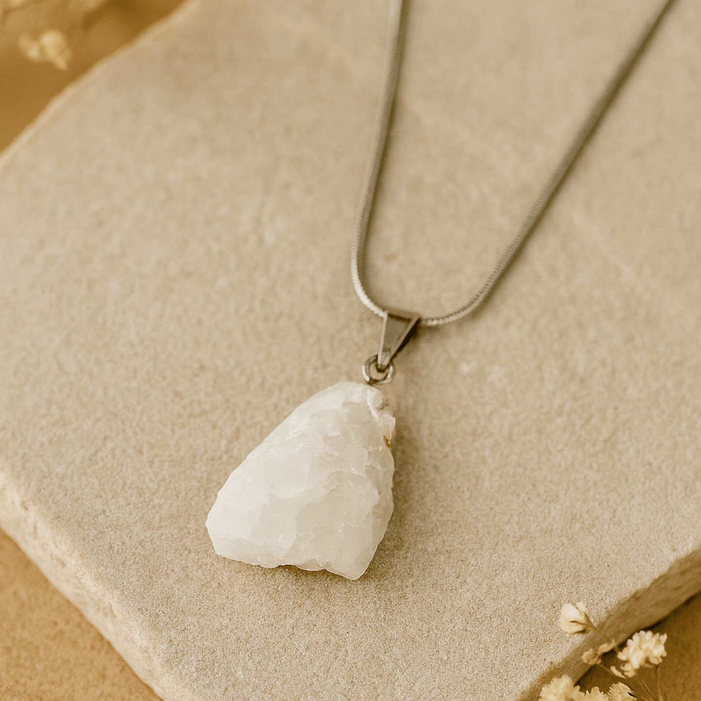 white moonstone rough pendant on a snake chain necklace on a beige stone and beige background a white flowers in the corner.