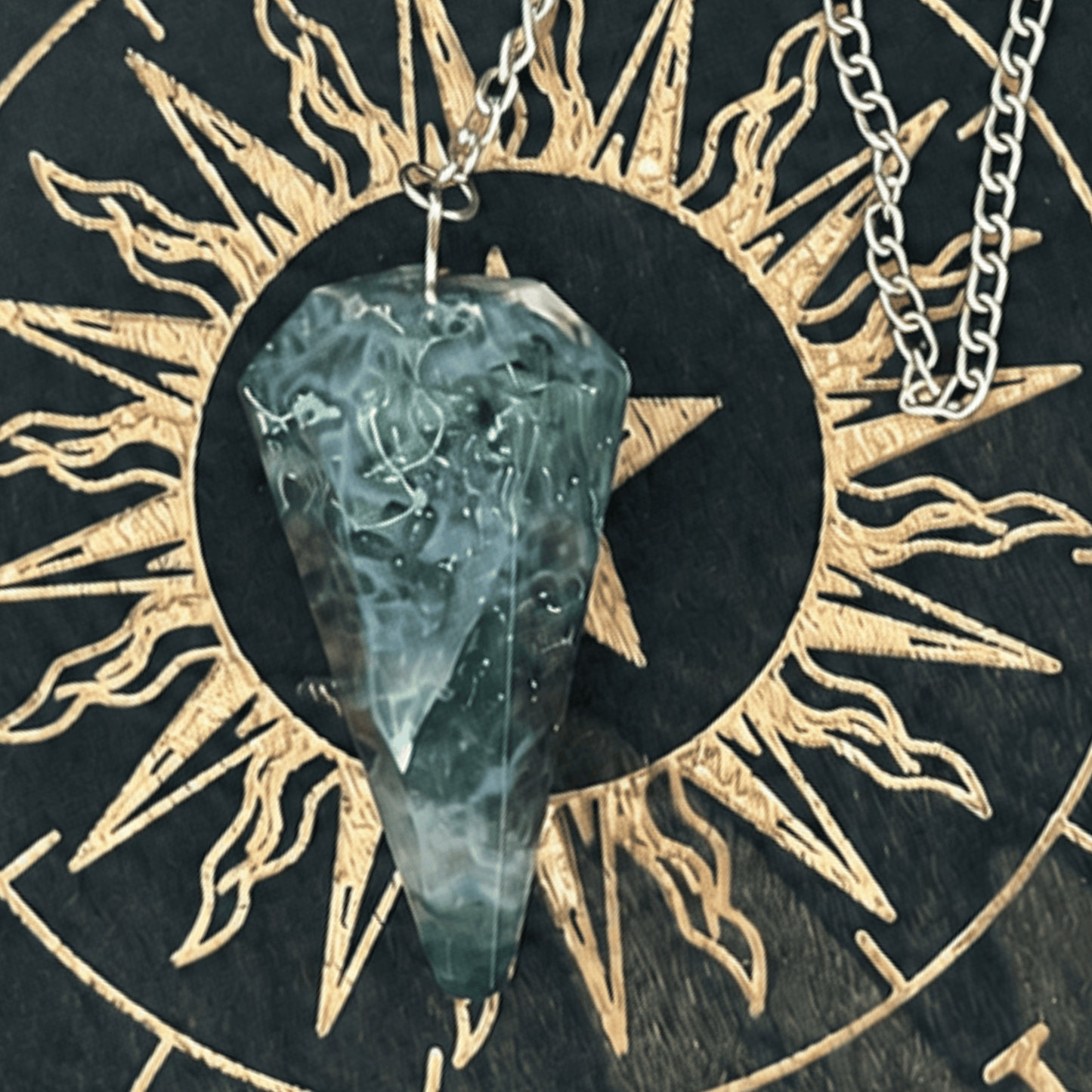 A silver chain necklace with a tumbled, pointed teal-green crystal pendant, displayed on a black and gold zodiac wheel background.