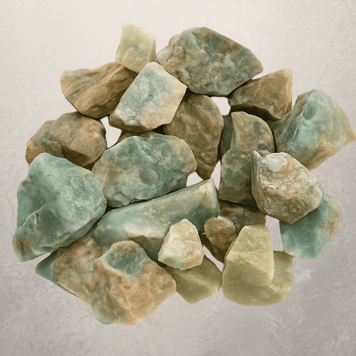 A pile of rough, raw amazonite crystals with natural earthy tan and light green hues, arranged on a textured light gray background.
