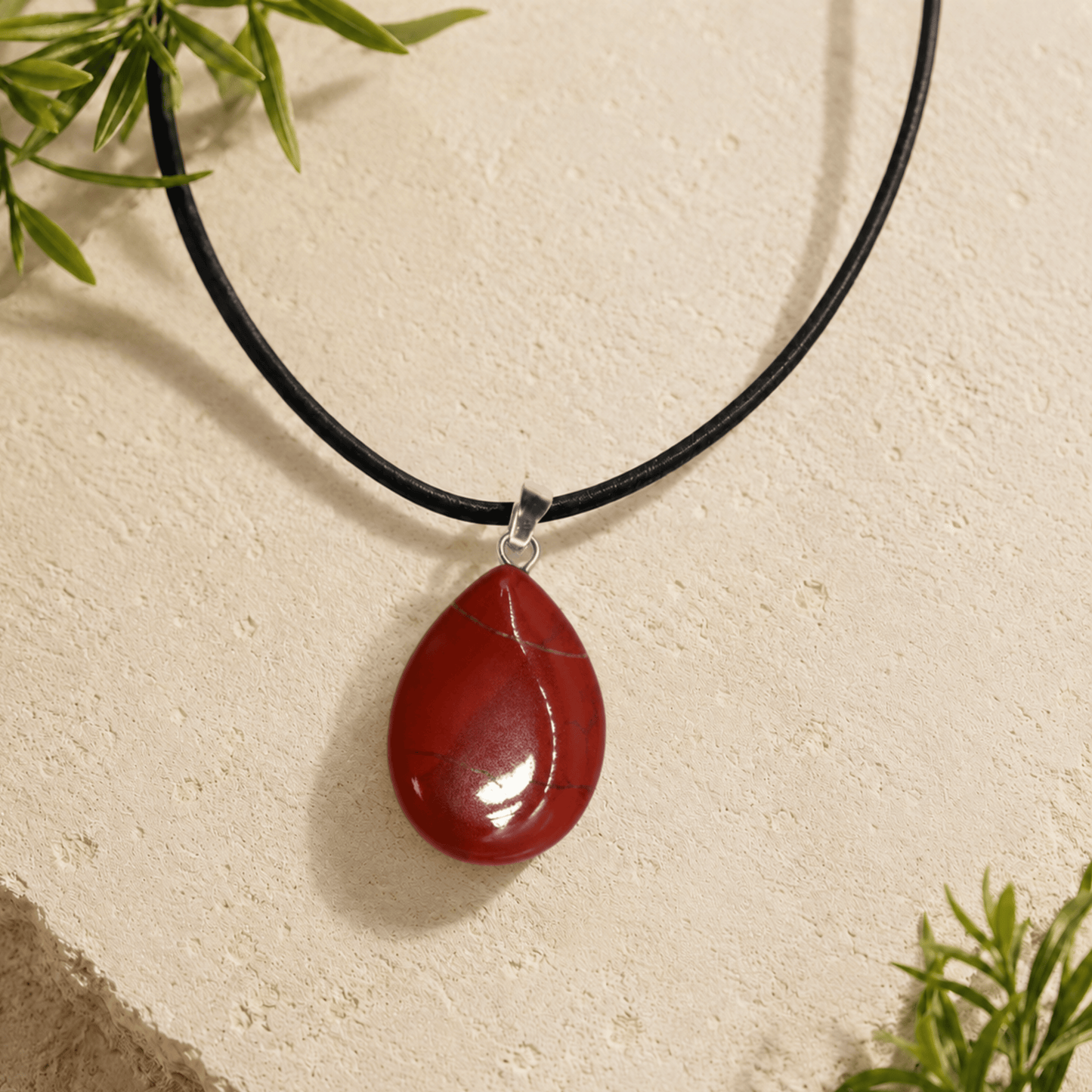Red teardrop pendant on a black cord against a beige background with green leaves.