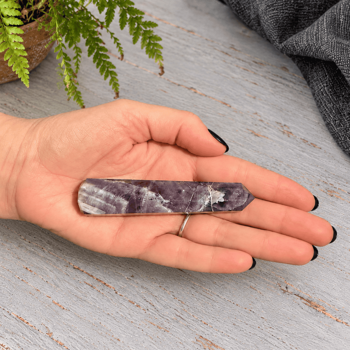 A polished purple amethyst crystal tower point resting on an open hand, with a silver ring on the ring finger, set against a rustic gray wooden background with a green fern in the corner.