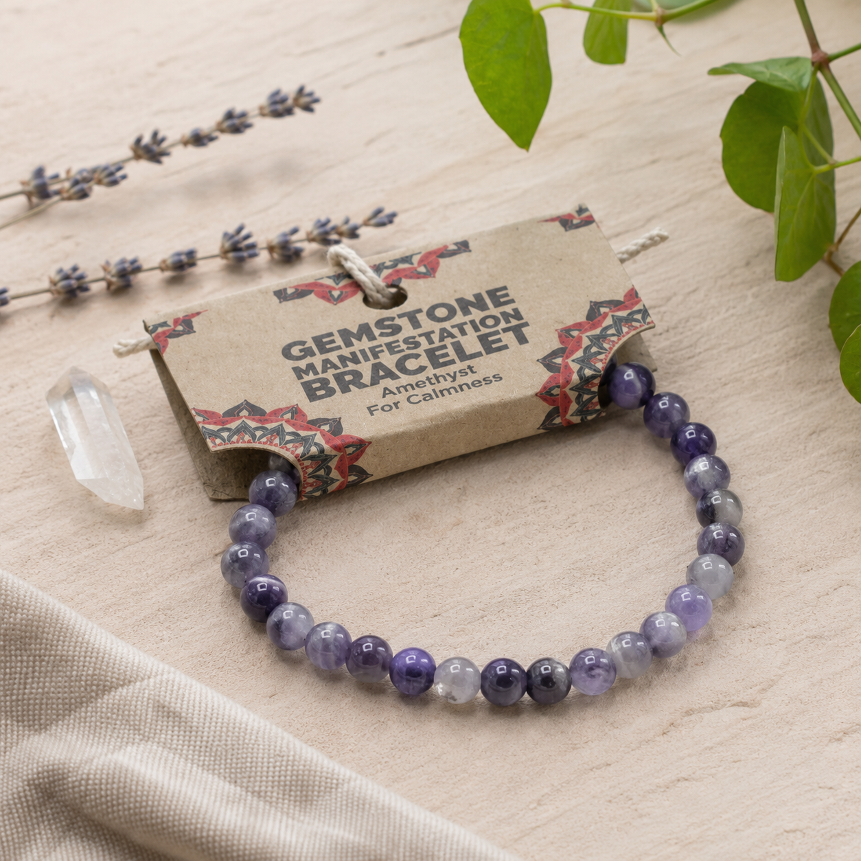 A beaded gemstone bracelet in shades of purple, lavender, and white, displayed on a kraft paper tag that reads 'GEMSTONE MANIFESTATION BRACELET Amethyst For Calmness.' The bracelet is styled on a light wood surface with a clear crystal, dried lavender, and green leaves.