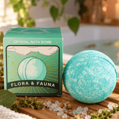 A turquoise crystal bath bomb sits beside its matching green and yellow illustrated box on a wooden surface, with bath salts and leaves nearby in a softly blurred bathroom setting.