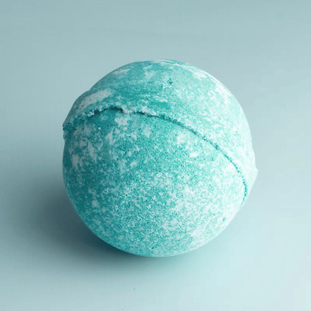 A round bath bomb with a textured, speckled mint green and white finish, sitting on a light blue-gray background.