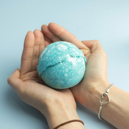 A person's hands holding a round, cracked turquoise bath bomb against a light blue background.