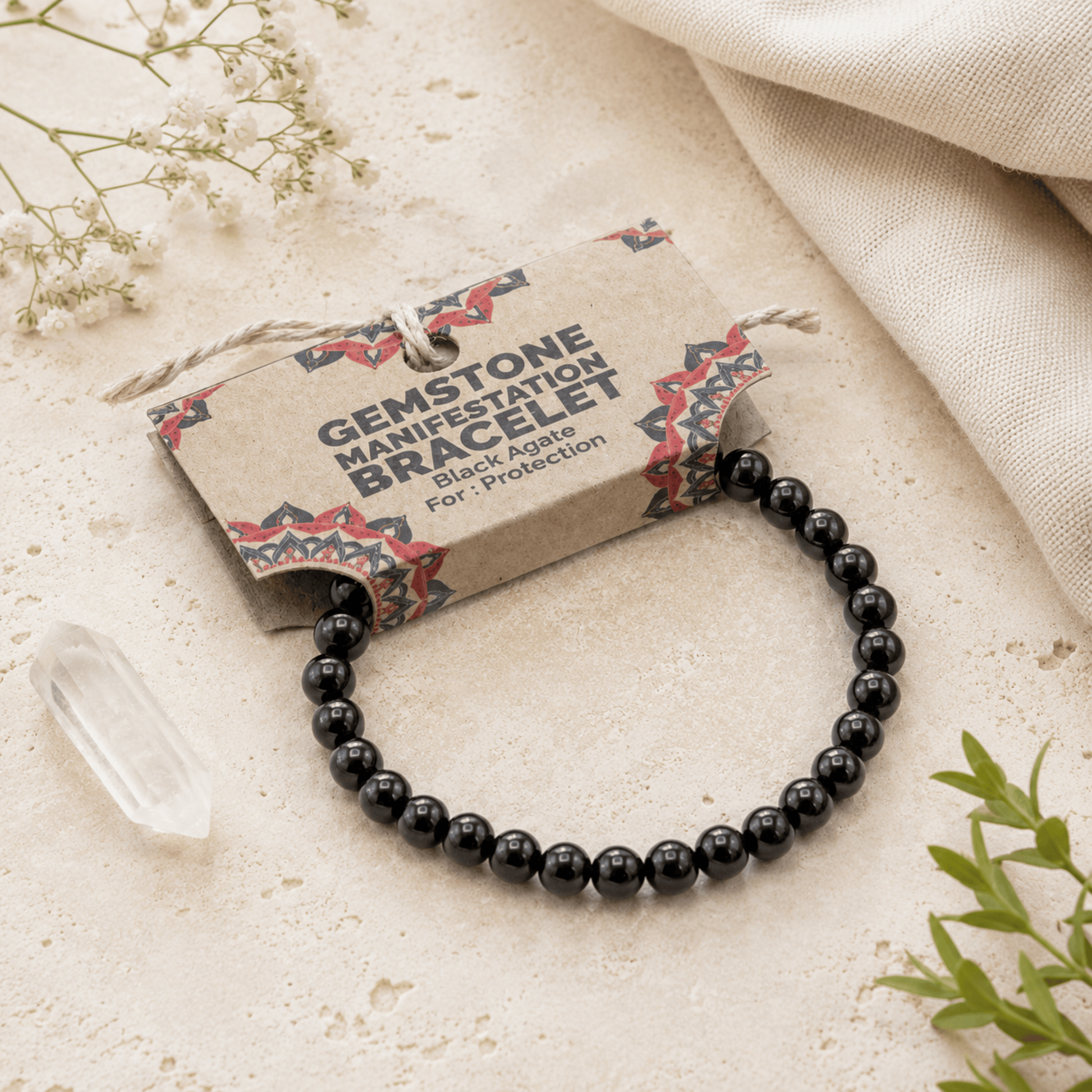 A black gemstone bracelet with polished round beads, attached to a kraft paper tag that reads 'GEMSTONE MANIFESTATION BRACELET Black Agate For Protection,' styled on a light stone surface with dried flowers, a clear crystal, and fabric.