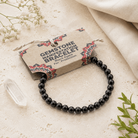 A black gemstone bracelet with polished round beads, attached to a kraft paper tag that reads 'GEMSTONE MANIFESTATION BRACELET Black Agate For Protection,' styled on a light stone surface with dried flowers, a clear crystal, and fabric.