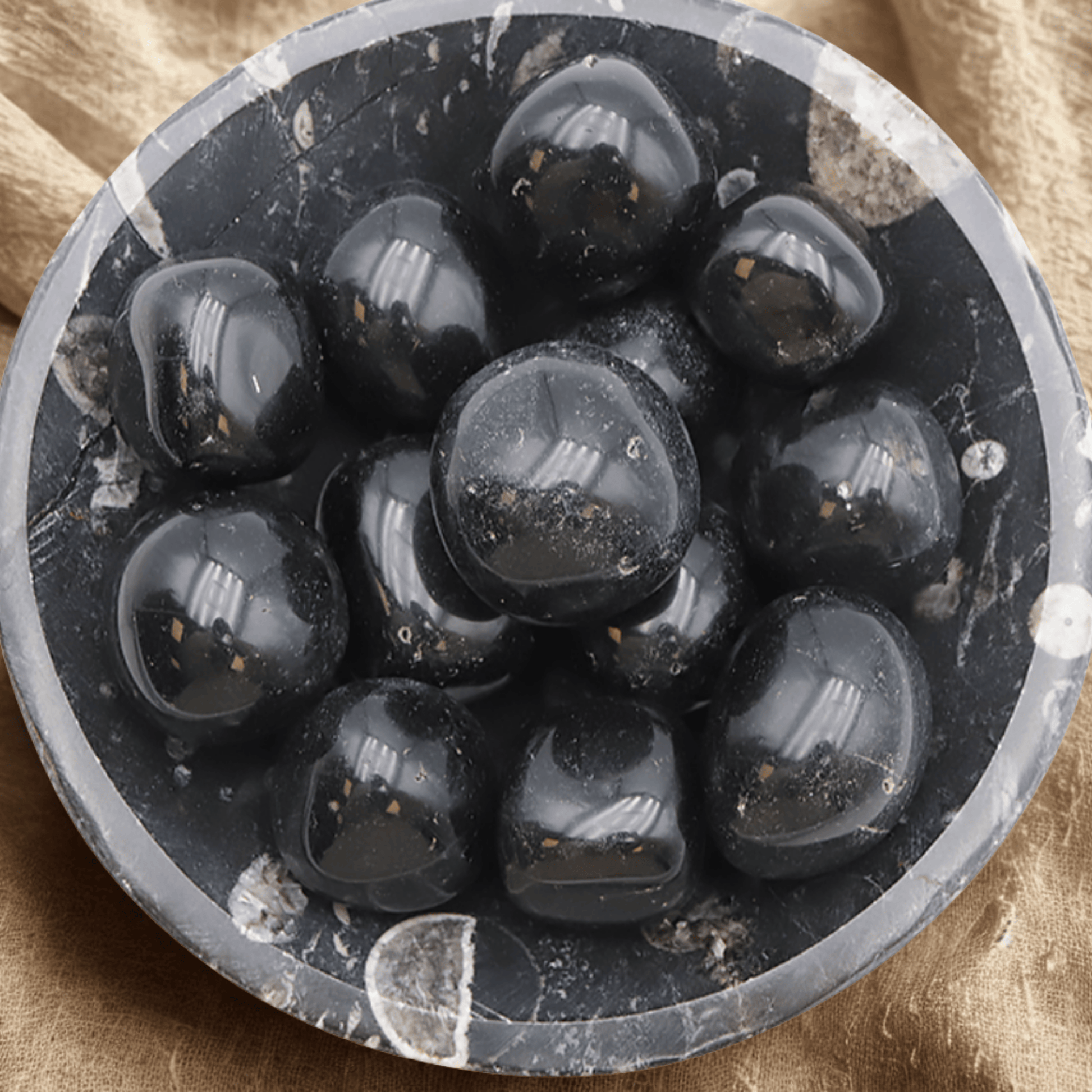 Close-up of a circular stone bowl with Black Obsidian tumblestones on a beige fabric background