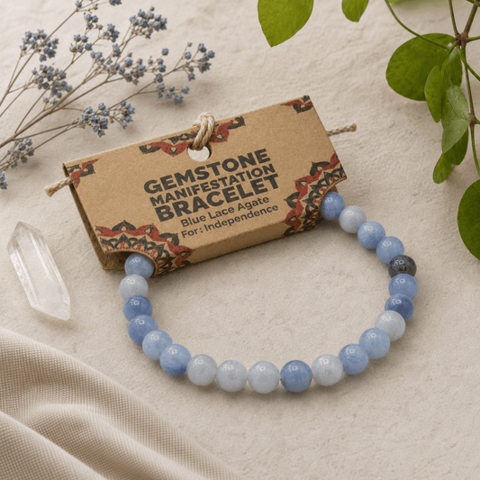 A beaded gemstone bracelet made of light blue, white, and dark blue agate beads, attached to a brown kraft paper card labeled 'GEMSTONE MANIFESTATION BRACELET Blue Lace Agate For: Independence.' The card is photographed on a beige fabric surface with dried blue flowers, green leaves, and a clear crystal point around it.