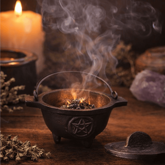 Small black cauldron with a carved pentagram in a circle on the front of it and herbs burning inside it with the lid laying next to it on a wooden table and herbs and crystal and a candle blurred in the background.