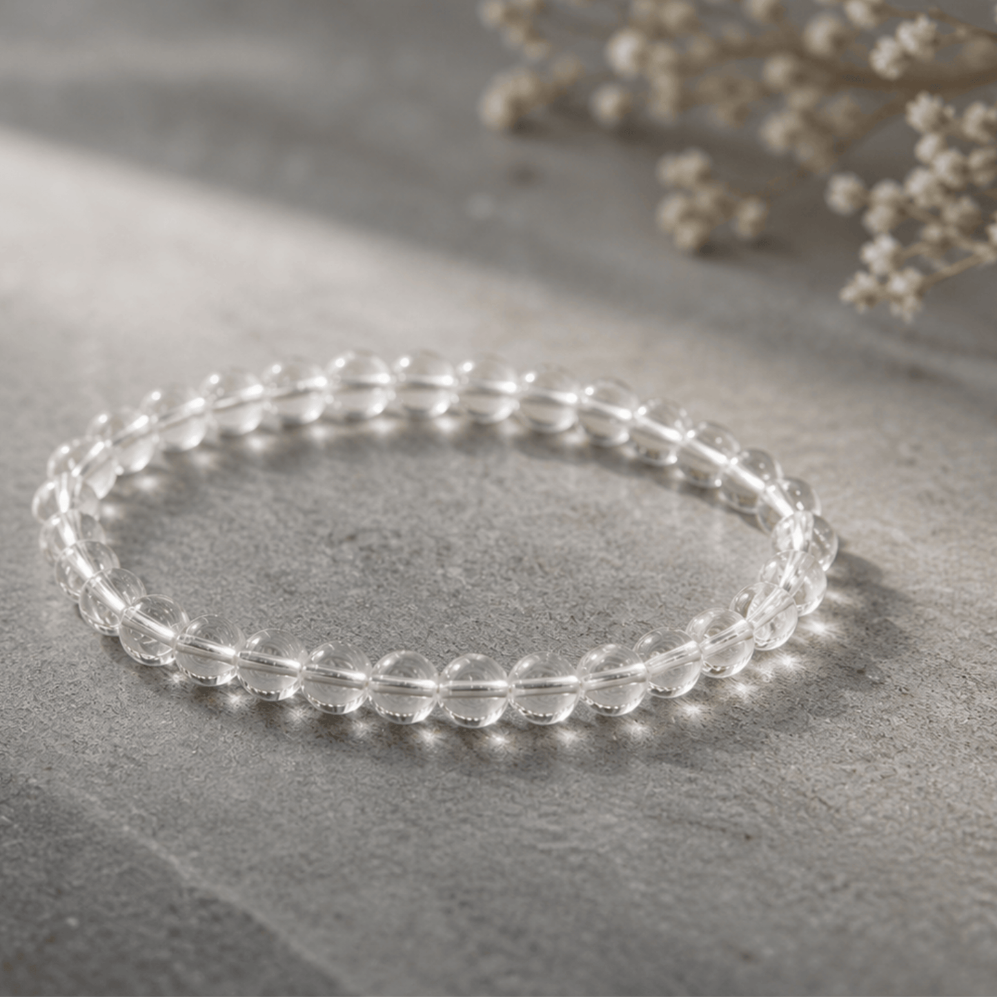 Clear beaded bracelet on a gray surface with light-colored flowers in the background