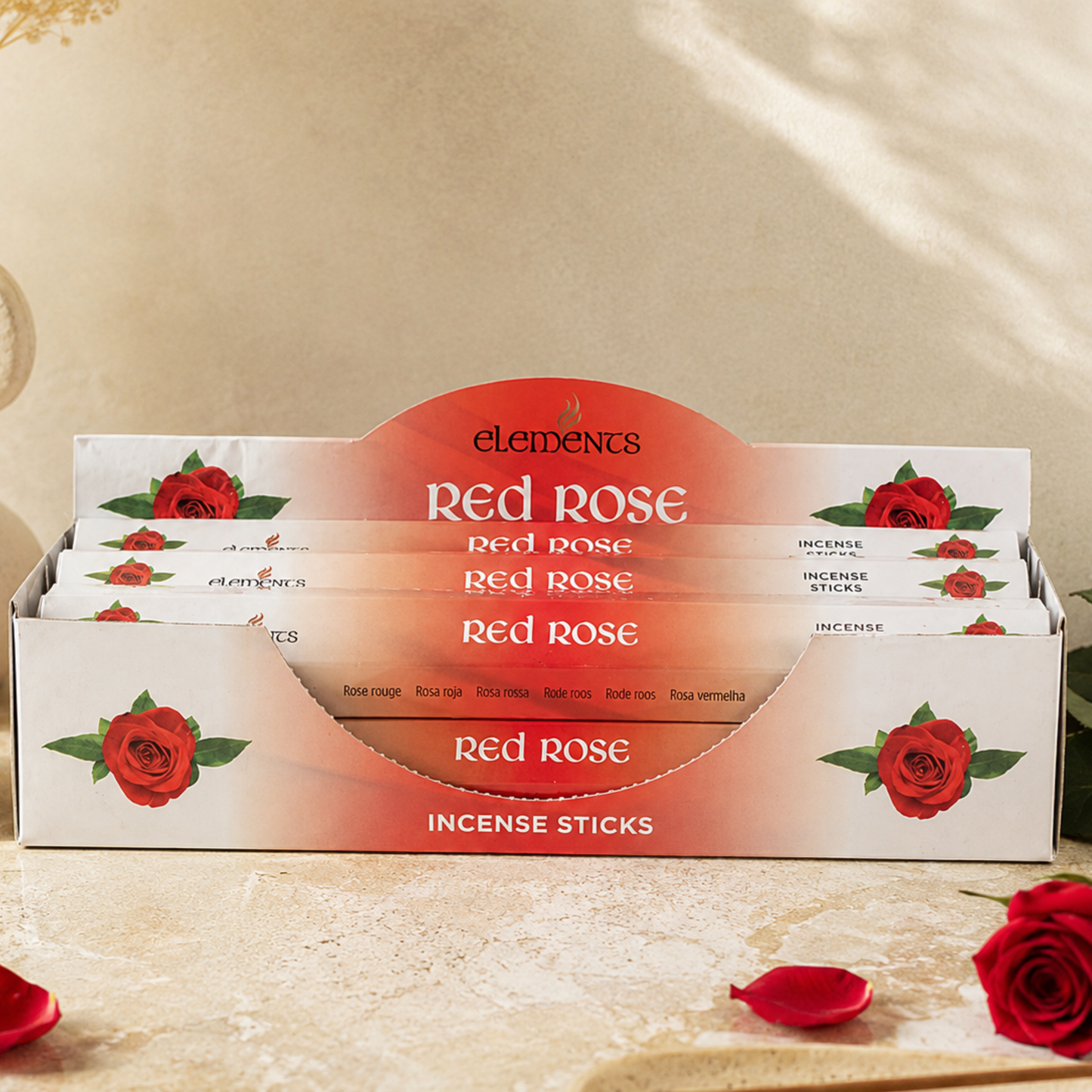 A display box of Elements Red Rose Incense Sticks, featuring white packaging decorated with red roses and a prominent red-orange arch label. The box is styled on a textured beige surface with scattered rose petals and a single red rose in the foreground.