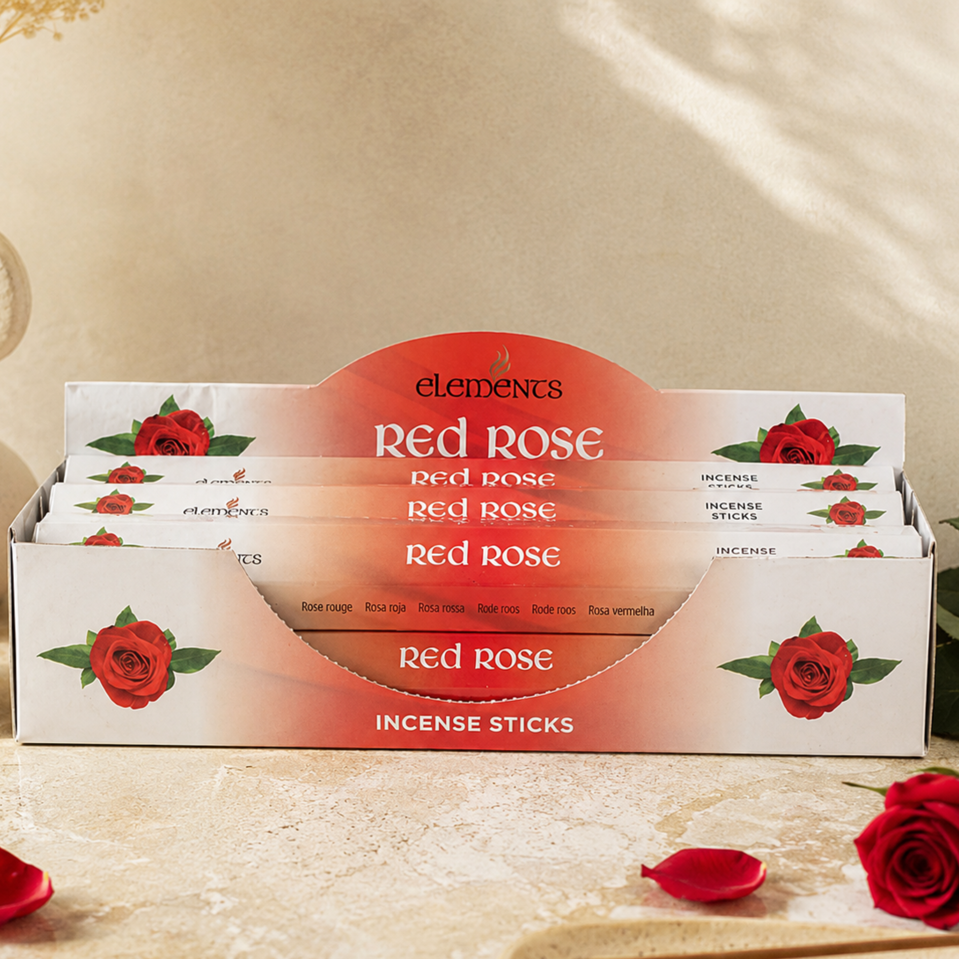 A display box of Elements Red Rose Incense Sticks, featuring white packaging decorated with red roses and a prominent red-orange arch label. The box is styled on a textured beige surface with scattered rose petals and a single red rose in the foreground.