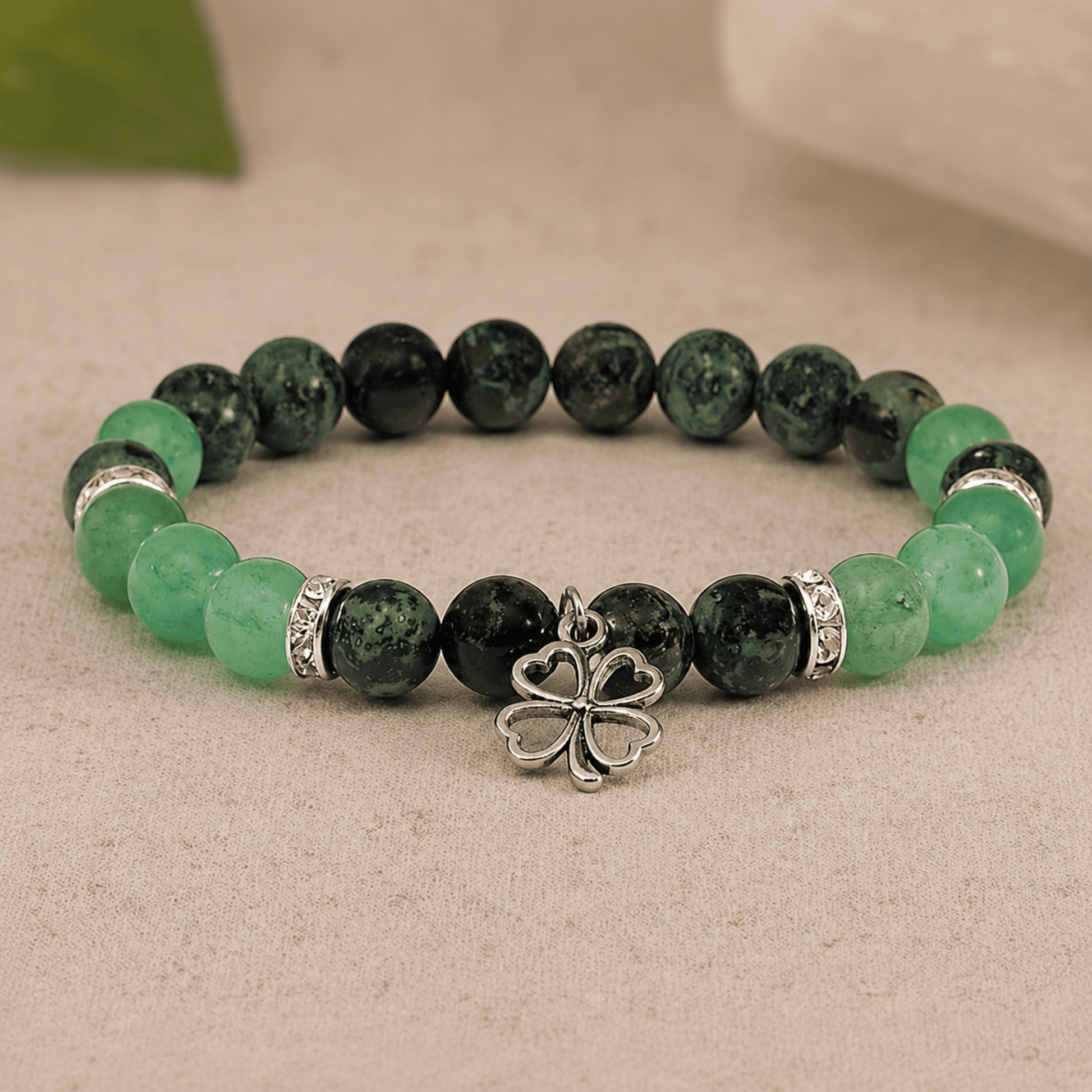 A beaded stretch bracelet featuring dark green marbled beads, light green translucent beads, silver spacer rings, and a central silver four-leaf clover charm, displayed on a neutral textured surface.