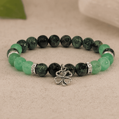 A beaded stretch bracelet featuring dark green marbled beads, light green translucent beads, silver spacer rings, and a central silver four-leaf clover charm, displayed on a neutral textured surface.