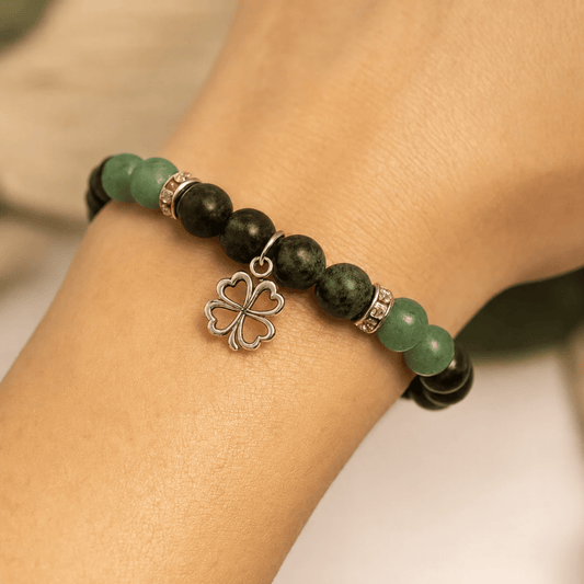A close-up of a beaded stretch bracelet worn on a wrist. The bracelet features dark green and black round beads, accented with silver spacer rings and a silver four-leaf clover charm.