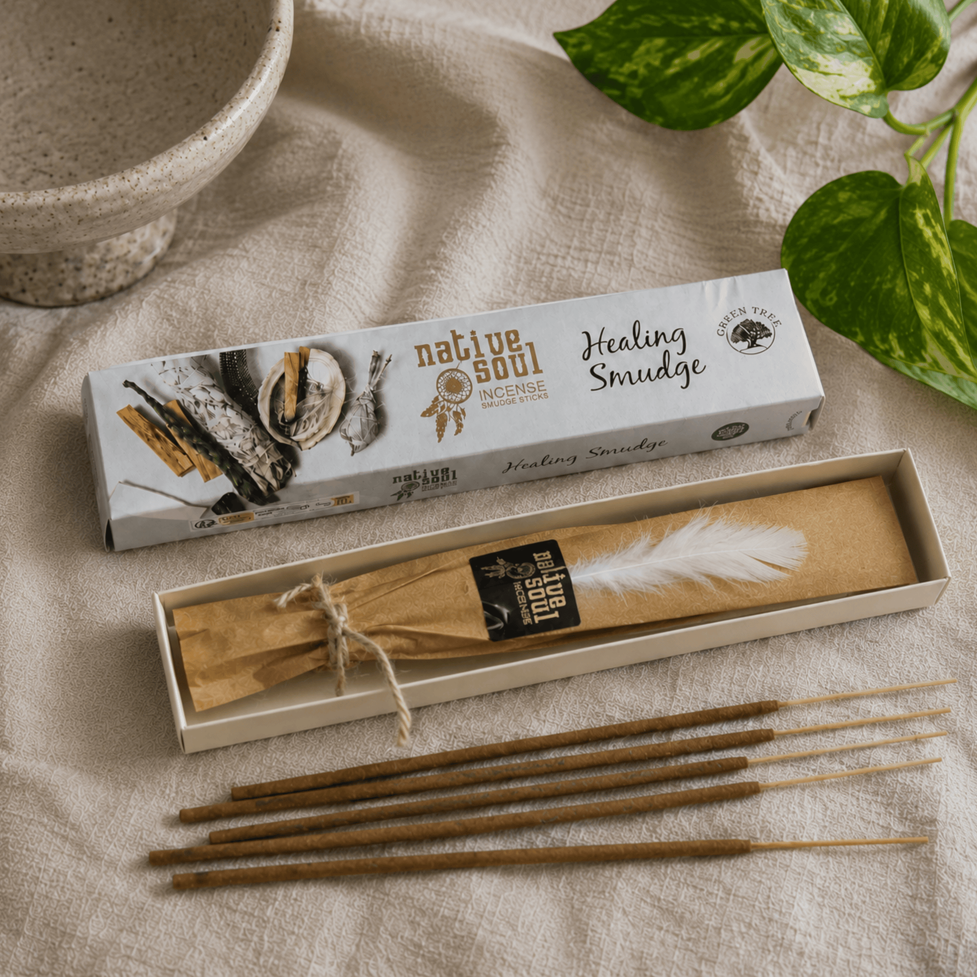 Native Soul Healing Smudge box set with a white and black box, an open box containing a brown paper-wrapped bundle with a white feather, several brown wooden snuff sticks, and a beige fabric background with green plant leaves.