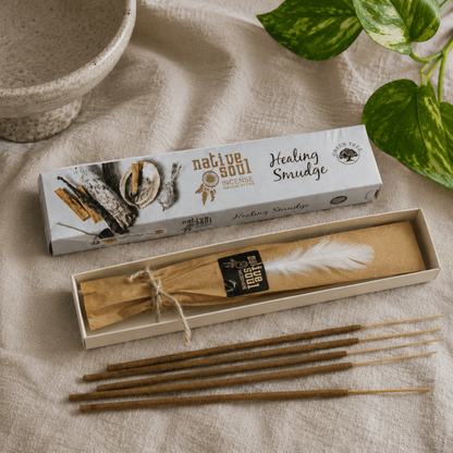 Native Soul Healing Smudge box set with a white and black box, an open box containing a brown paper-wrapped bundle with a white feather, several brown wooden snuff sticks, and a beige fabric background with green plant leaves.
