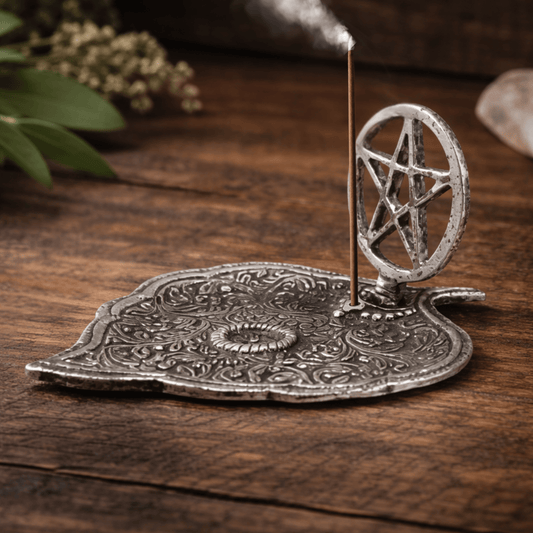 Silver incense holder tray shaped as a leaf and a pentagram in a circle on the one end on a wooden table and an incense stick burning.