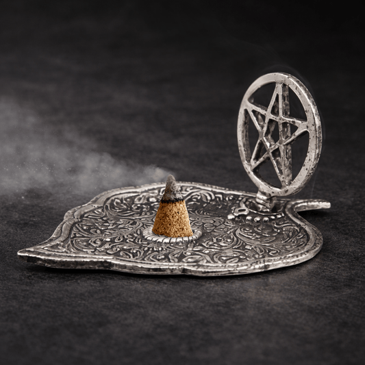 Silver incense holder tray shaped as a leaf and a pentagram in a circle on the one end on a black background and an incense cone burning