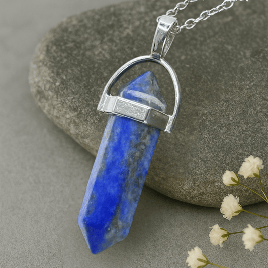 Lapis Lazuli Blue crystal pendant with a zinc alloy horse shoe shape attachment for the silver chain laying against  a stone and flowers in the background