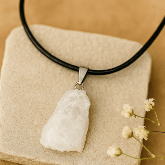 white moonstone rough pendant on a black cord necklace on a beige stone and beige background a white flowers in the corner.
