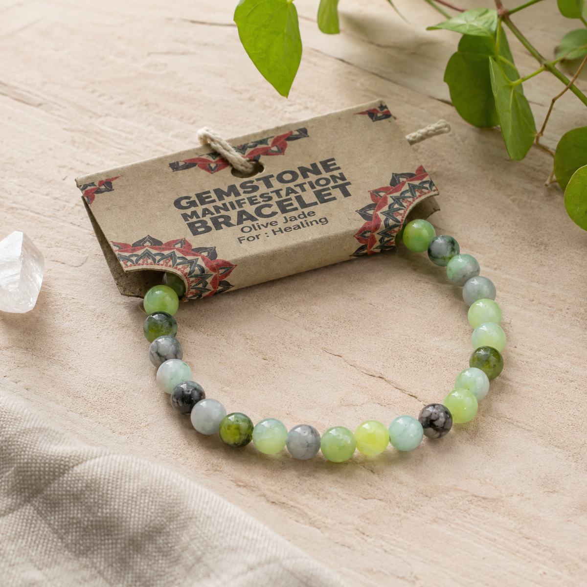 A beaded gemstone bracelet with olive jade, light green, gray, and white beads, displayed on its kraft paper hang tag with a small clear crystal and green leaves in the background.