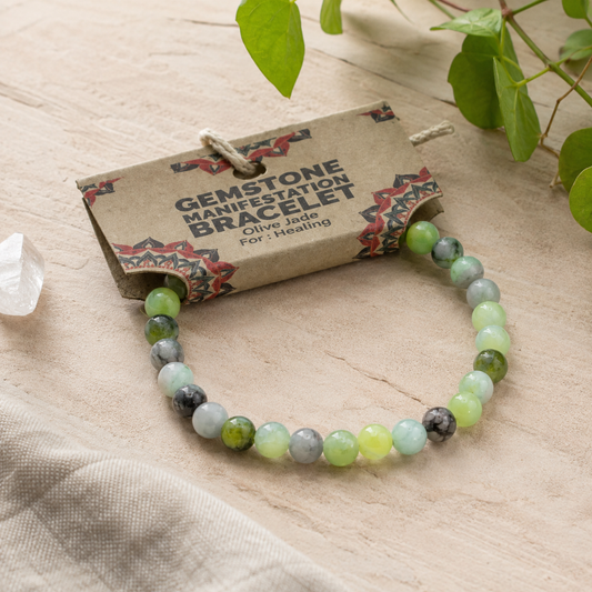 A beaded gemstone bracelet with olive jade, light green, gray, and white beads, displayed on its kraft paper hang tag with a small clear crystal and green leaves in the background.