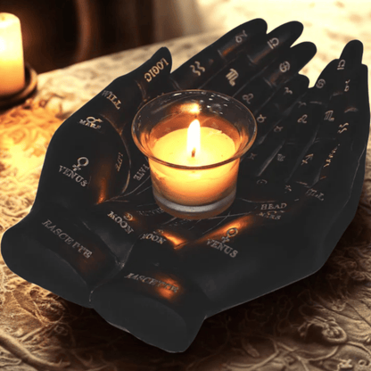 Black resin hands with zodiac symbols holding a lit candle on a textured surface.
