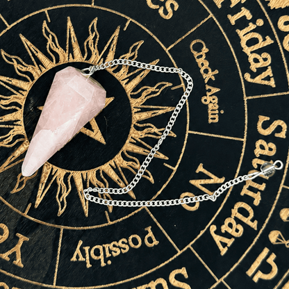 A silver chain pendulum with a pink rose quartz crystal facet point, laid out on a black and gold astrology wheel.