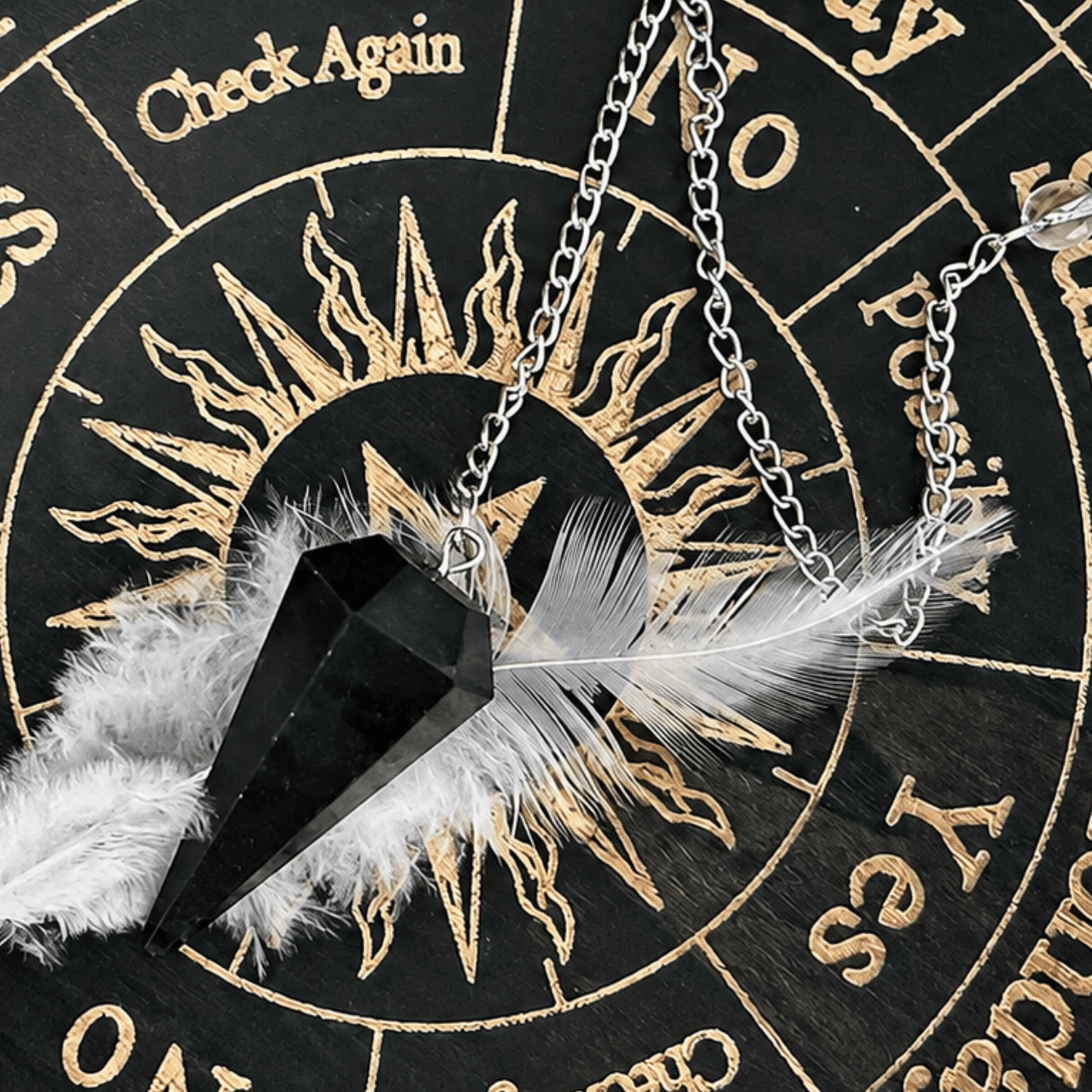A silver chain necklace with a black crystal point pendant, decorated with a white feather and fluffy white feather, displayed on a black and gold astrological wheel.