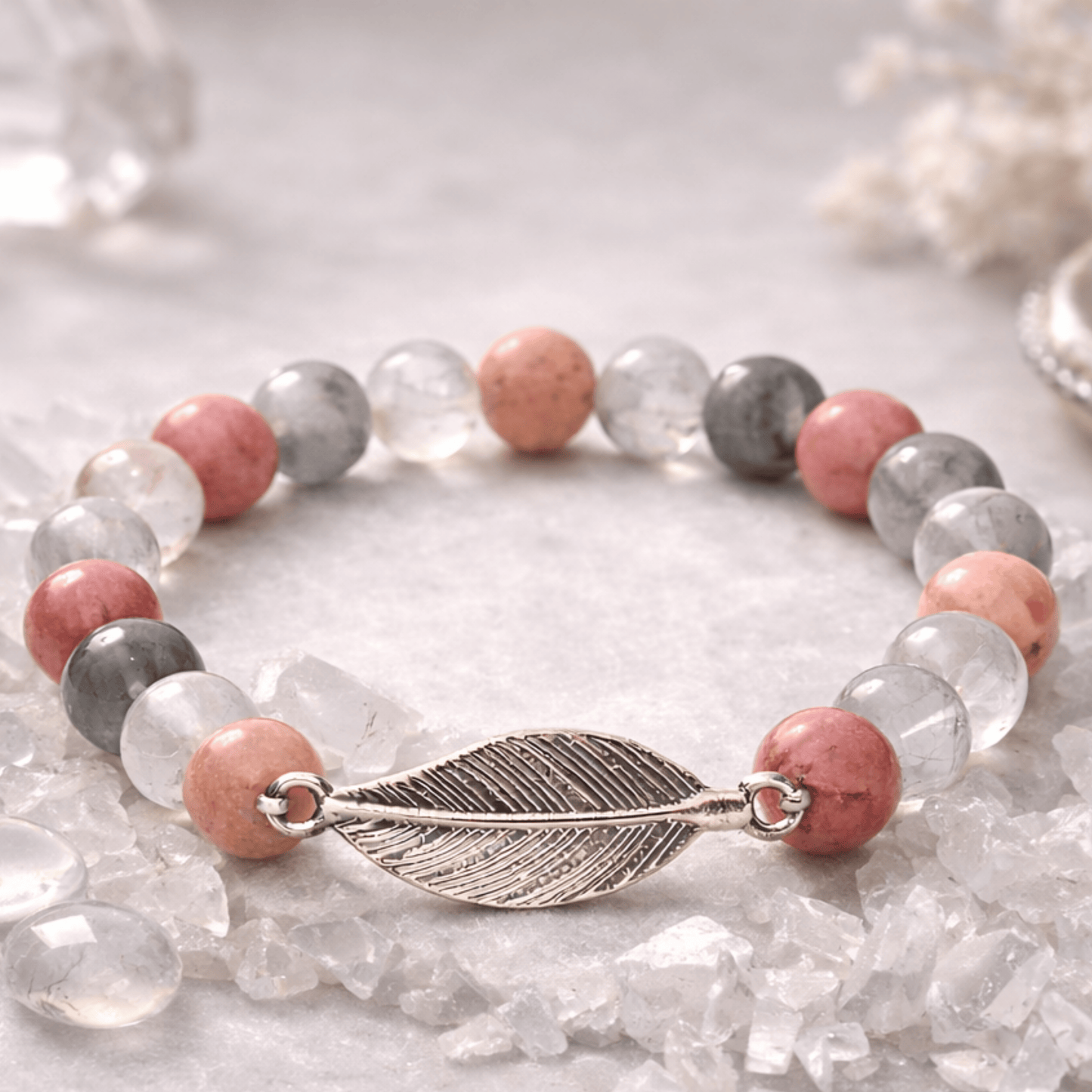 Rhodonite & Grey Quartz Mala Bracelet with Feather Charm on a beige background with rock crystlchips laying around it.