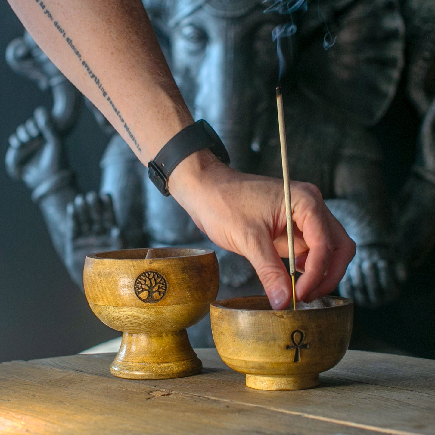 Hand placing an incense stick into a wooden bowl with a tree of life symbol, against a blurred background.
