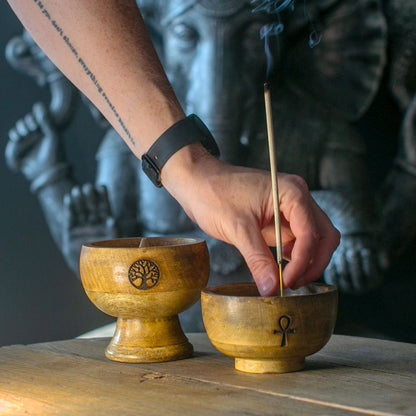 Hand placing an incense stick into a wooden bowl with a tree of life symbol, against a blurred background.