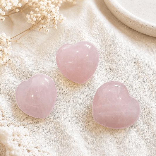 Three smooth, polished pink heart-shaped stones arranged on a textured cream fabric with dried white flowers and a ceramic dish edge in the background.