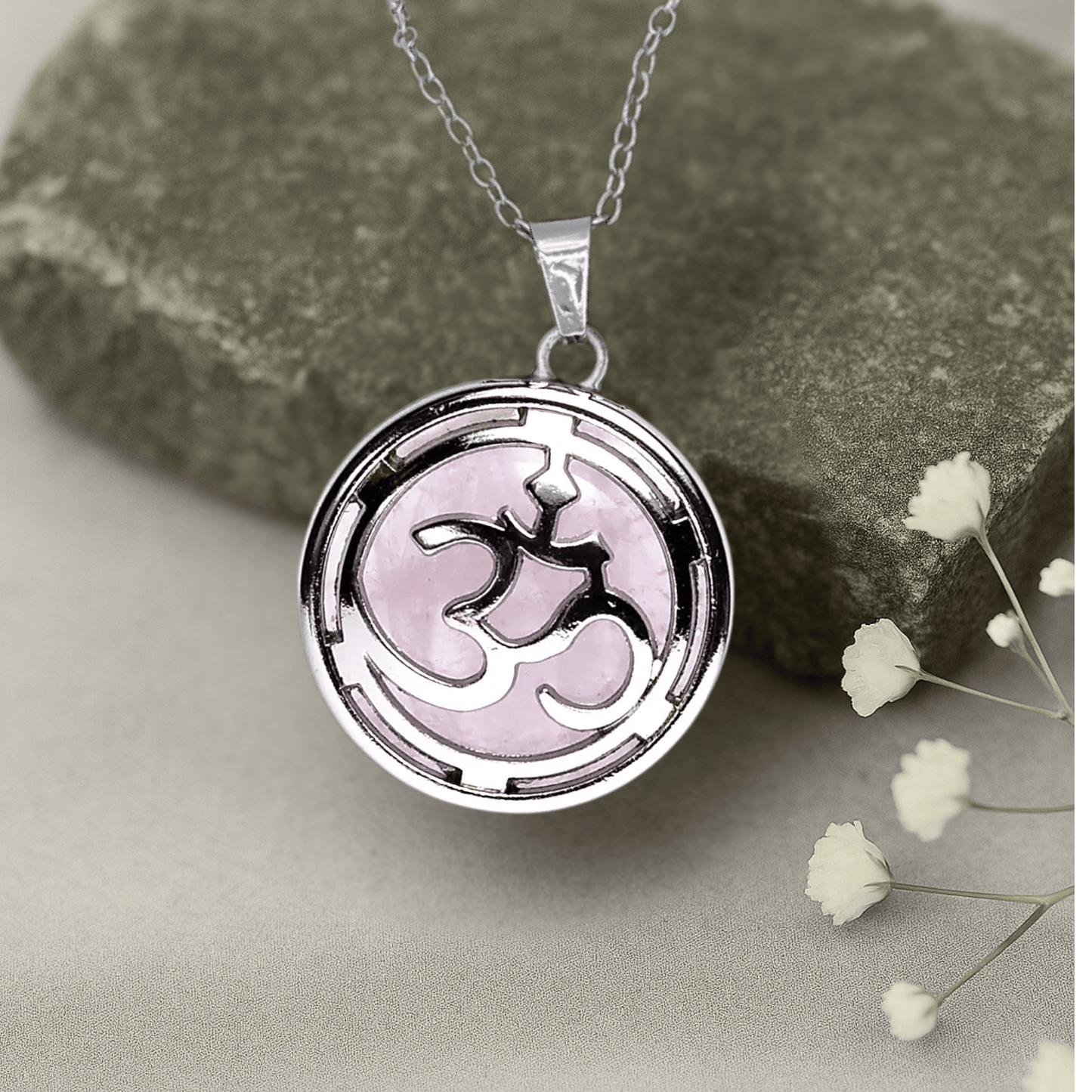 Silver necklace with a pink rose quartz enclosed in a pendant with the ohm symbol.*
The clasp on top is made of copper and the base holding the stone is zinc alloy  on a stone surface with small white flowers.