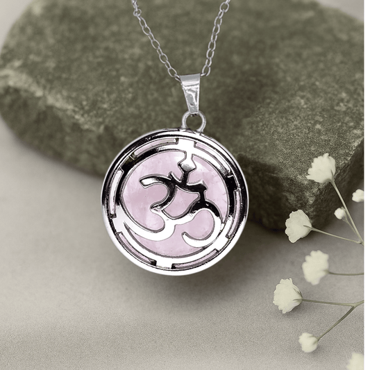 Silver necklace with a pink rose quartz enclosed in a pendant with the ohm symbol.*
The clasp on top is made of copper and the base holding the stone is zinc alloy  on a stone surface with small white flowers.