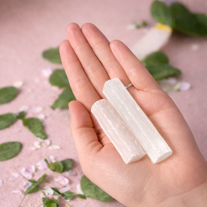 Hand holding two white crystal sticks with a natural background of leaves and flowers.