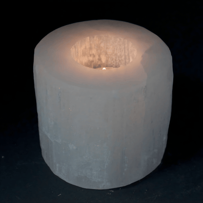 White Selenite Crystal candle holder with a lit tea light candle inside the hole of the cylinder on a black background