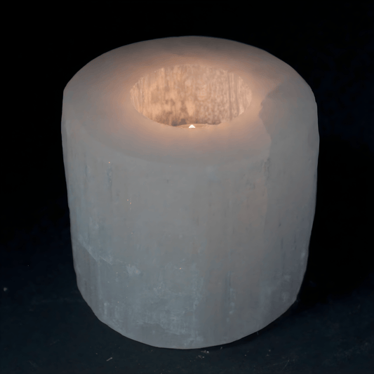 White Selenite Crystal candle holder with a lit tea light candle inside the hole of the cylinder on a black background