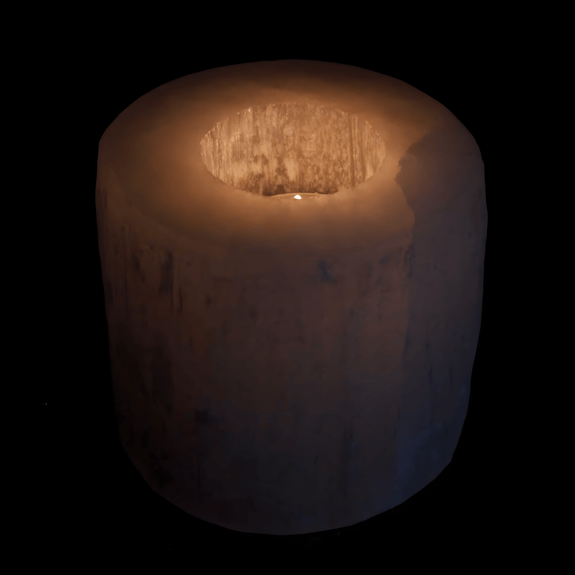 White Selenite Crystal candle holder with a lit tea light candle inside the hole of the cylinder . The cylinder glows with a warm glow on a black background