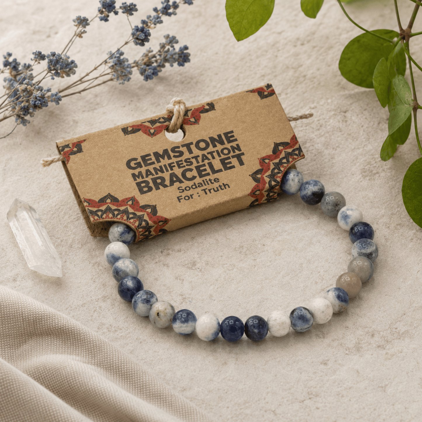 A gemstone manifestation bracelet made of blue, white, and gray agate-like beads, displayed on a kraft paper tag that reads 'GEMSTONE MANIFESTATION BRACELET' with 'Sodalis For: Truth' printed on it. The bracelet is styled on a neutral fabric surface with dried flowers, green leaves, and a clear crystal point.