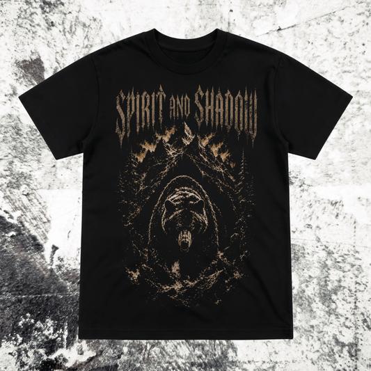 A black short-sleeve graphic t-shirt featuring the text 'SPIRIT AND SHADOW' in a distressed metallic gold font above a dark, fiery hooded figure illustration, displayed flat against a textured gray and white background.