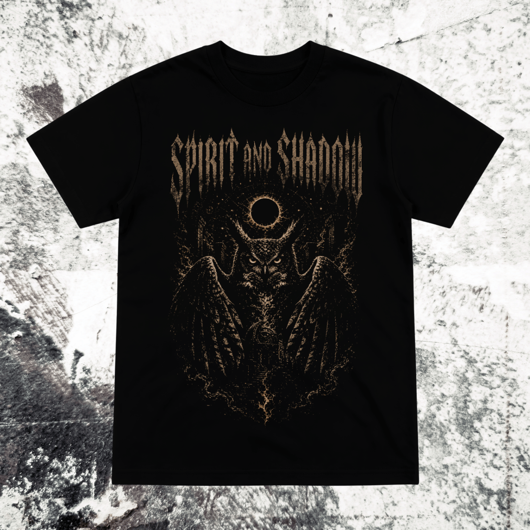 A black short-sleeve graphic t-shirt featuring a metallic gold gothic-style print of a winged skull with a circular symbol above its head and the text 'SPIRIT AND SHADOW' across the chest, displayed flat against a textured gray and white background.