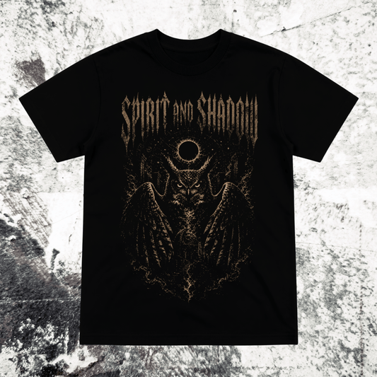 A black short-sleeve graphic t-shirt featuring a metallic gold gothic-style print of a winged skull with a circular symbol above its head and the text 'SPIRIT AND SHADOW' across the chest, displayed flat against a textured gray and white background.