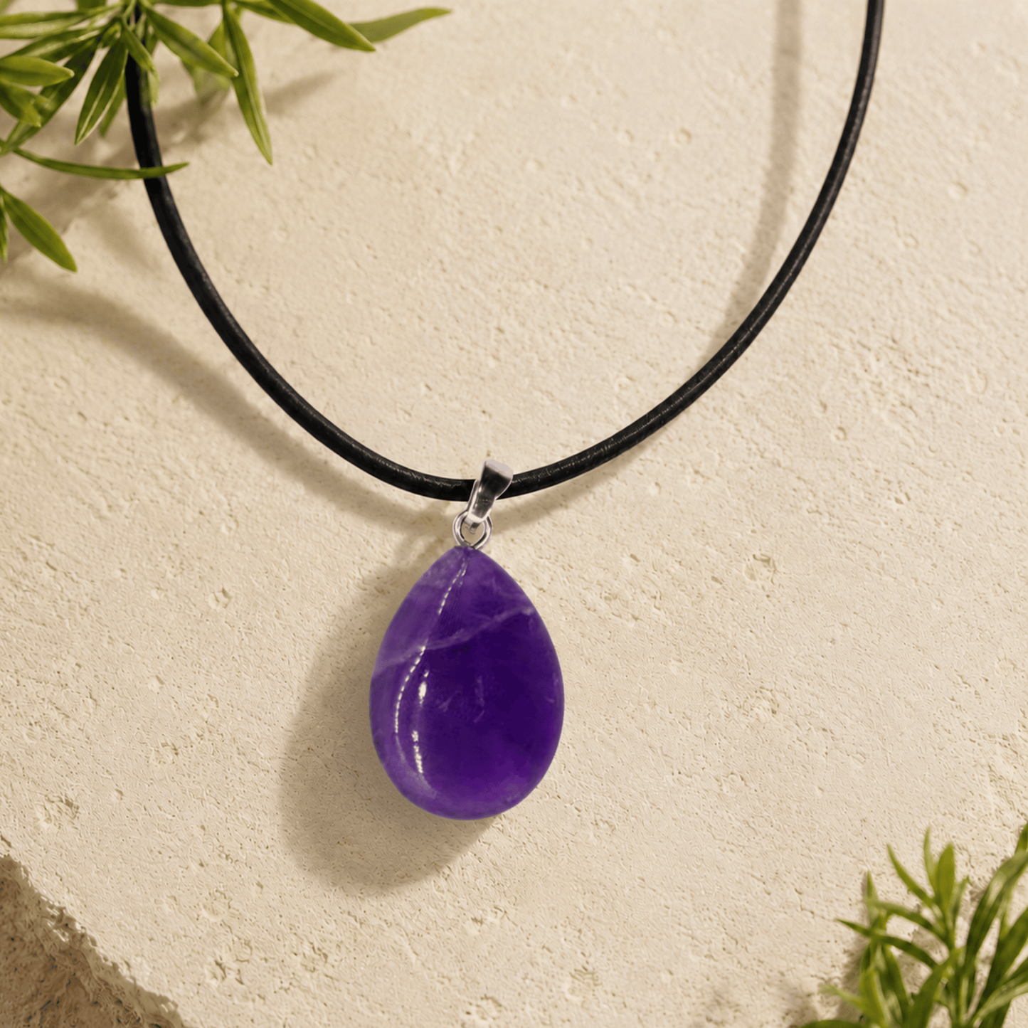 Purple teardrop pendant on a black cord against a beige background with green leaves.