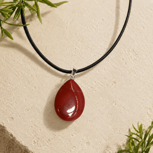 Red teardrop pendant on a black cord against a beige background with green leaves.